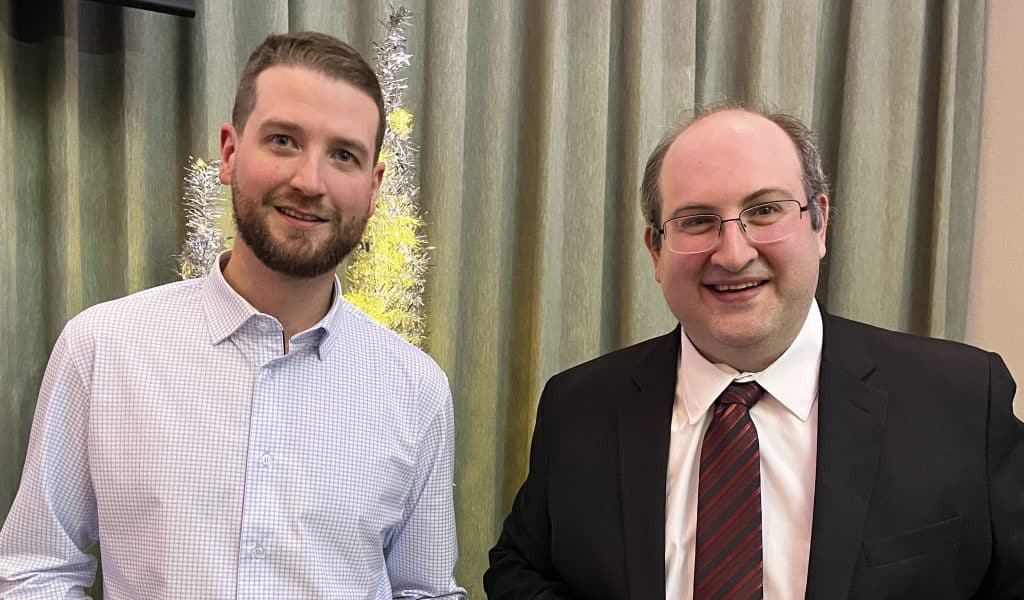 Young professional man in suit and casual shoulders at a corporate event, standing next to a decorative tree in a warm indoor setting.