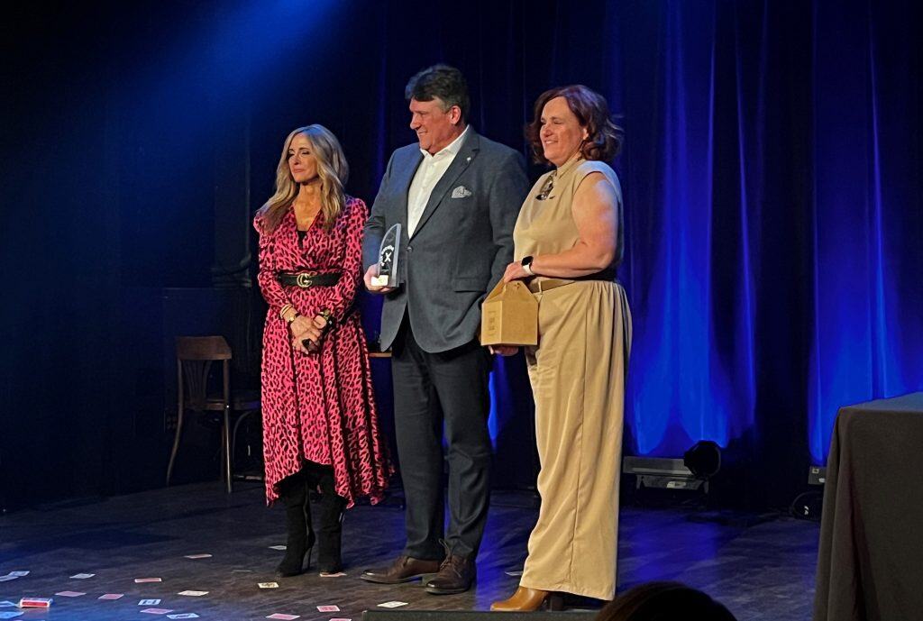 Homme recevant un prix lors d'une cérémonie avec deux femmes sur scène, ambiance élégante et formelle.