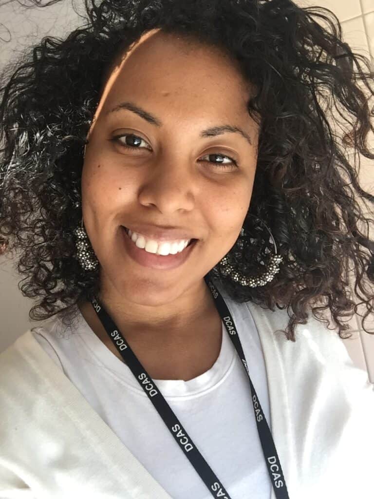Smiling woman with soft, healthy natural curls and statement earrings wearing a white top and work badge.