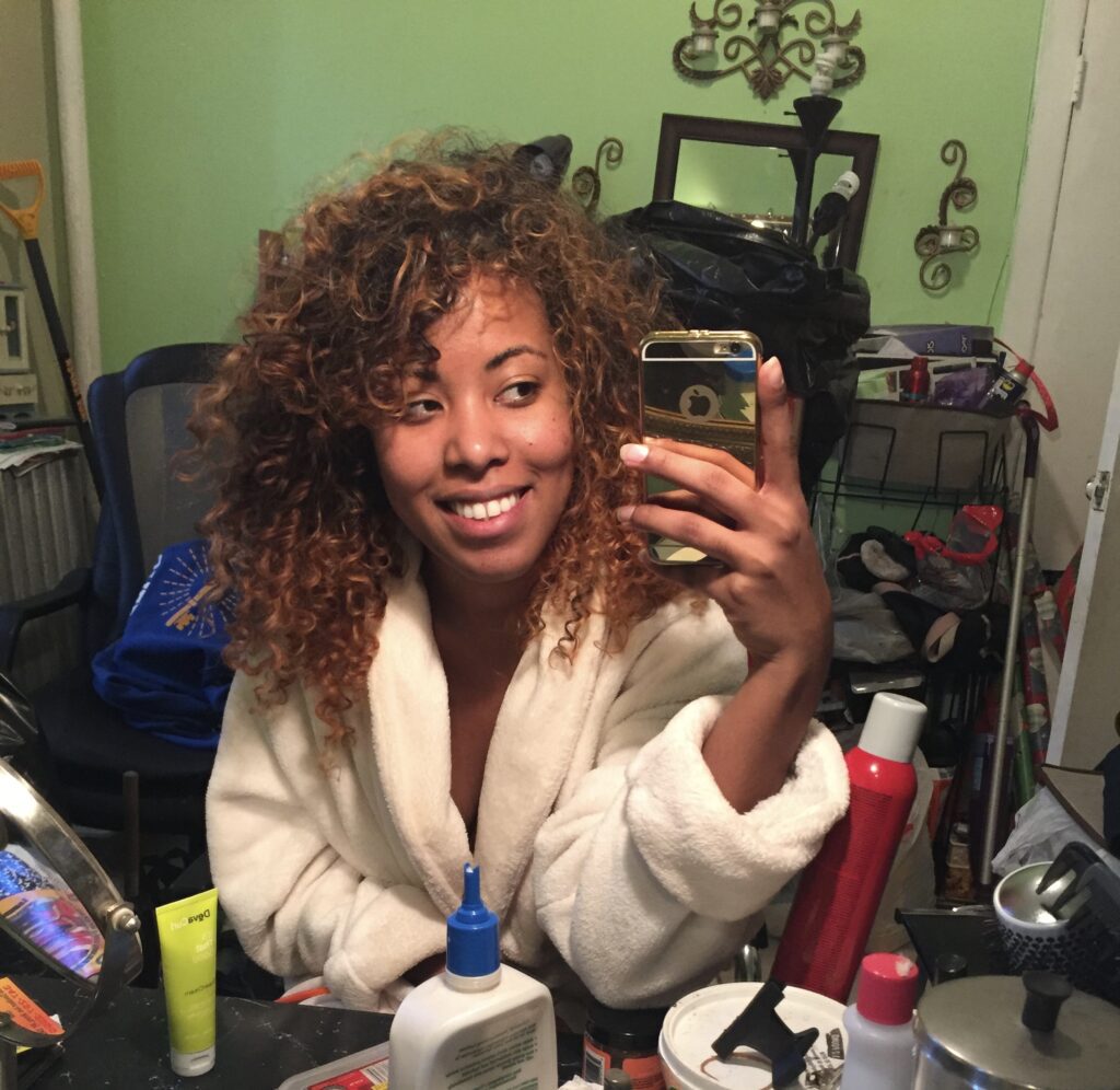 Smiling woman with defined curls and healthy natural hair taking a selfie at home, surrounded by curly hair care products and styling tools.