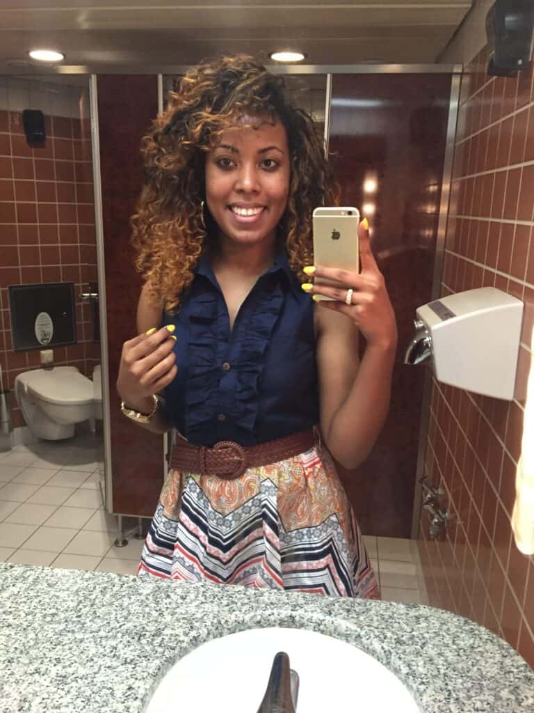 Woman with highlighted curls, wearing a navy blue crop top and patterned skirt, smiling in a bathroom mirror selfie.