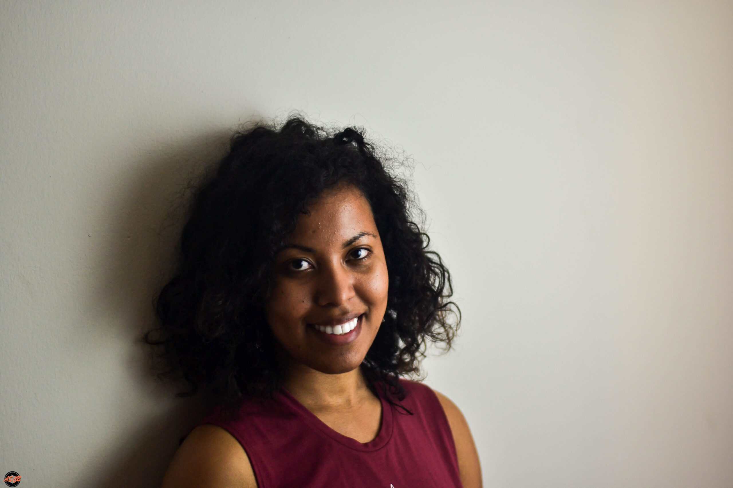 Woman with loosely defined curls and a serious expression, wearing a geometric tank top, posing against a neutral background.