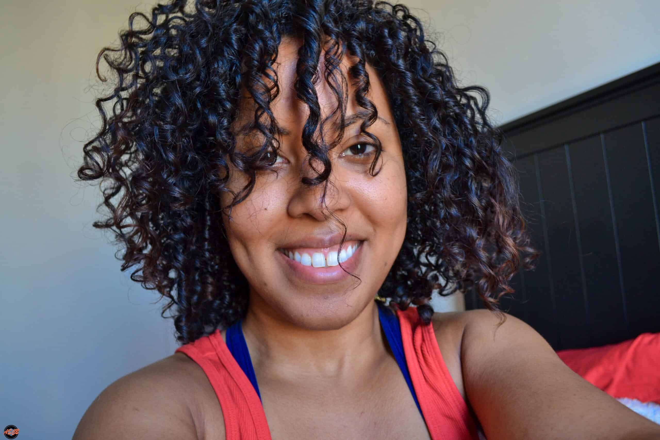 Close-up of a woman with defined, moisturized curls and minimal makeup, smiling in bright indoor light.