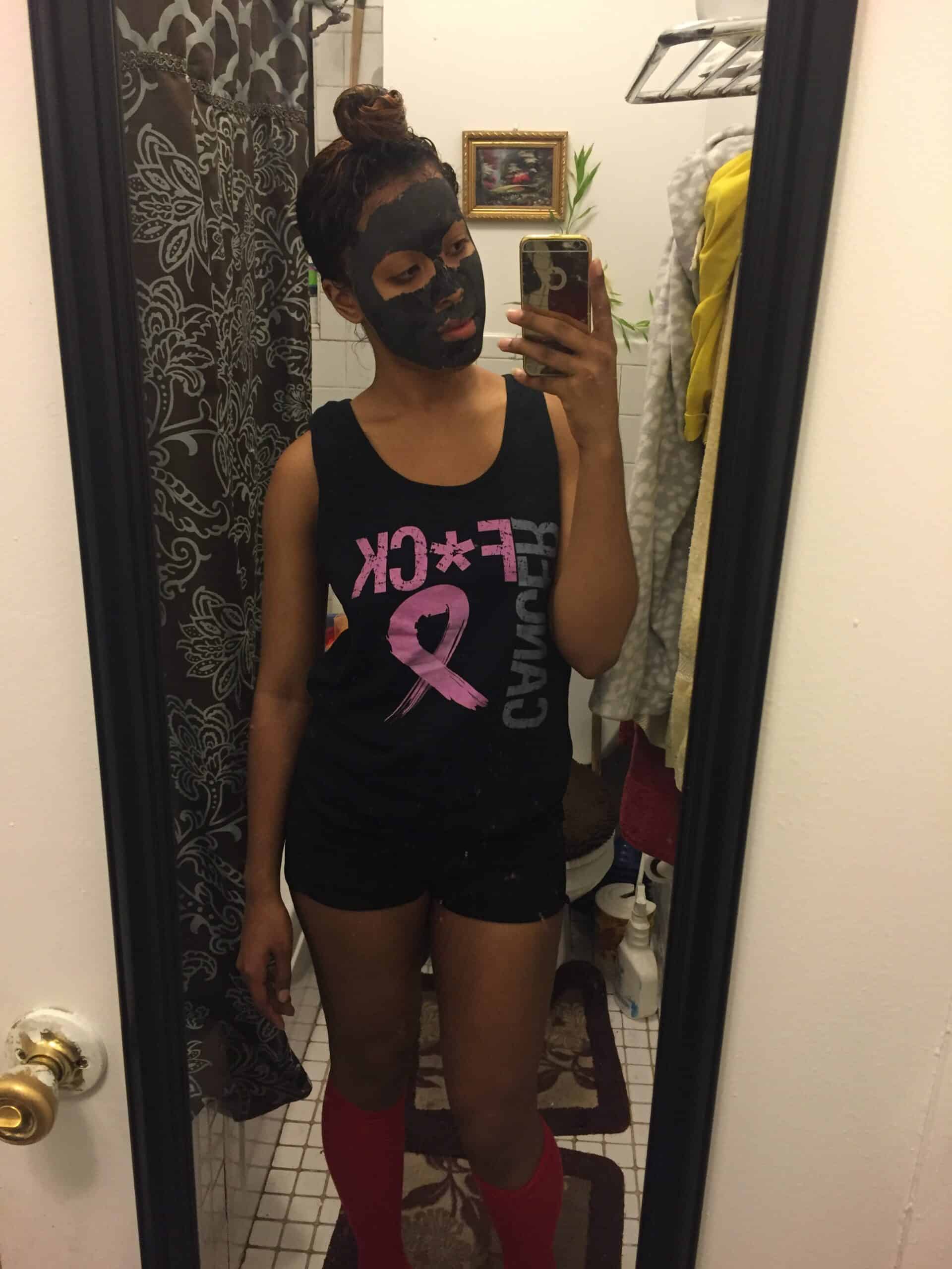 Woman taking a mirror selfie while wearing a black bentonite clay face mask, mid-treatment in a casual at-home setting.