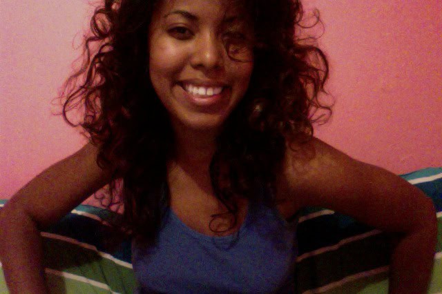 Smiling woman with voluminous natural curls sitting on a bed against a pink wall, wearing a blue tank top
