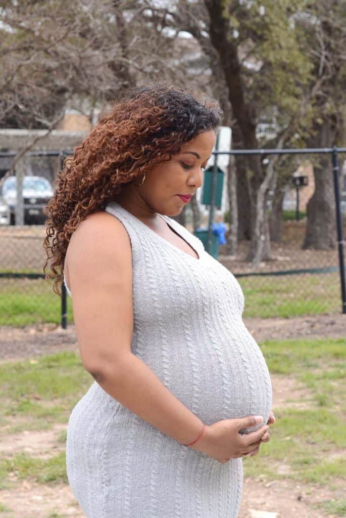 A close-up of a pregnant woman in a gray dress with her hands gently tucked under her baby bump, looking down lovingly at her belly during a home maternity photoshoot.