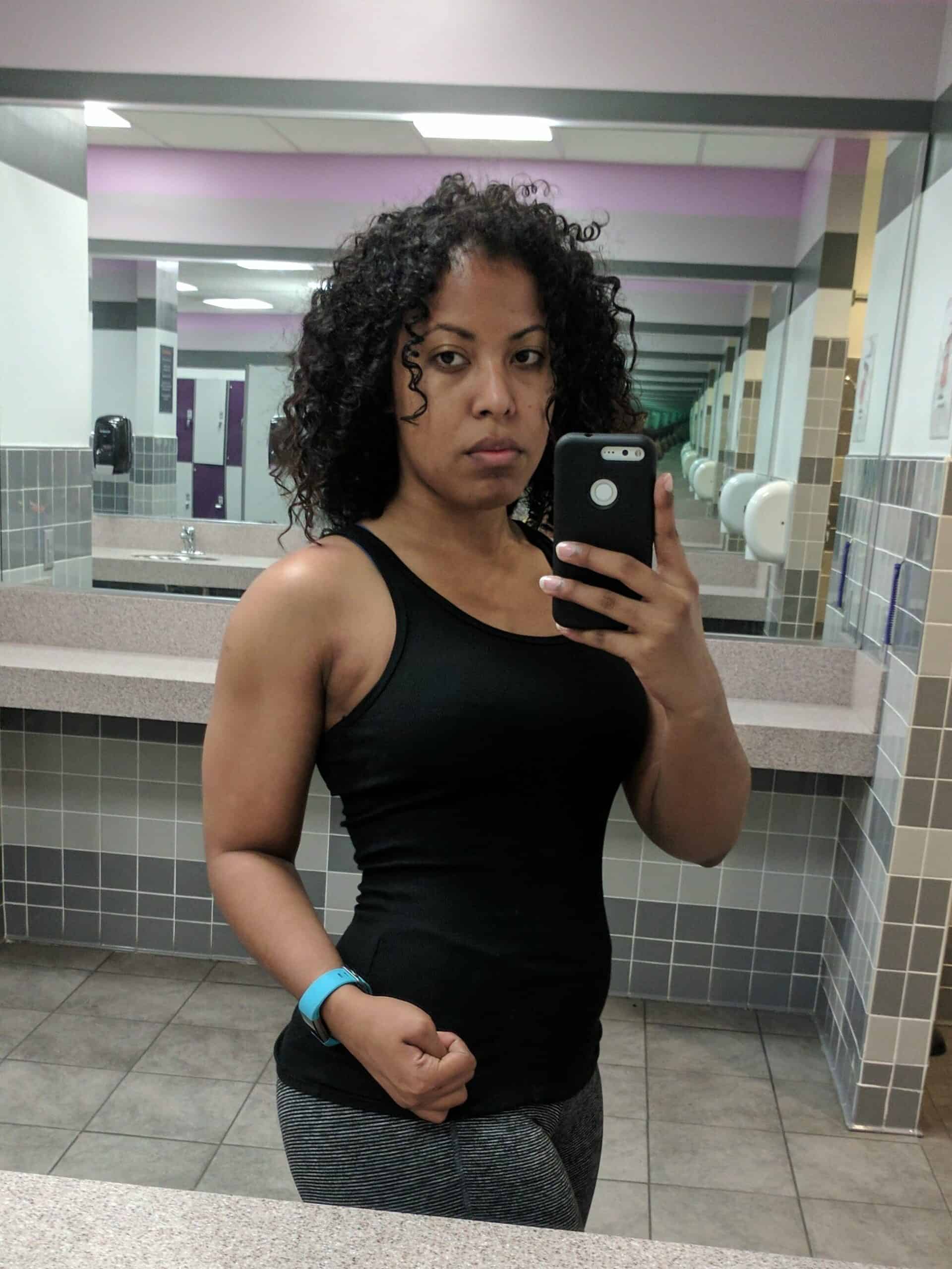 Woman taking a mirror selfie at the gym, flexing her arm and showing visible muscle definition while wearing a black tank top.
