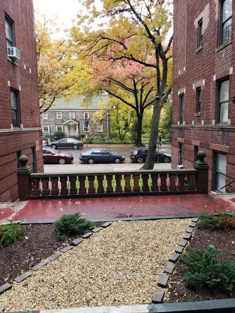 A peaceful brick apartment courtyard with a walking path, trees, and blooming plants during early fall.