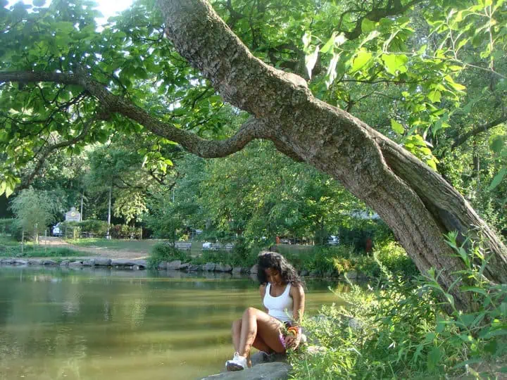 Woman sitting by a tranquil pond under a large leaning tree in a quiet area of Van Cortlandt Park, surrounded by summer greenery.