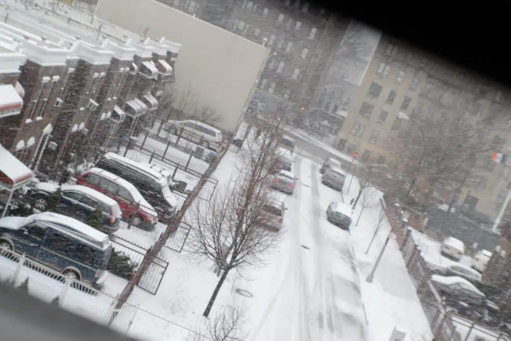 A residential Bronx street blanketed in snow, with rows of parked cars and brownstone buildings during winter.