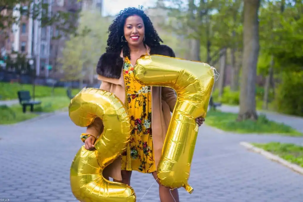 Woman holding gold “27” birthday balloons, standing on a tree-lined sidewalk and smiling on a sunny day.
