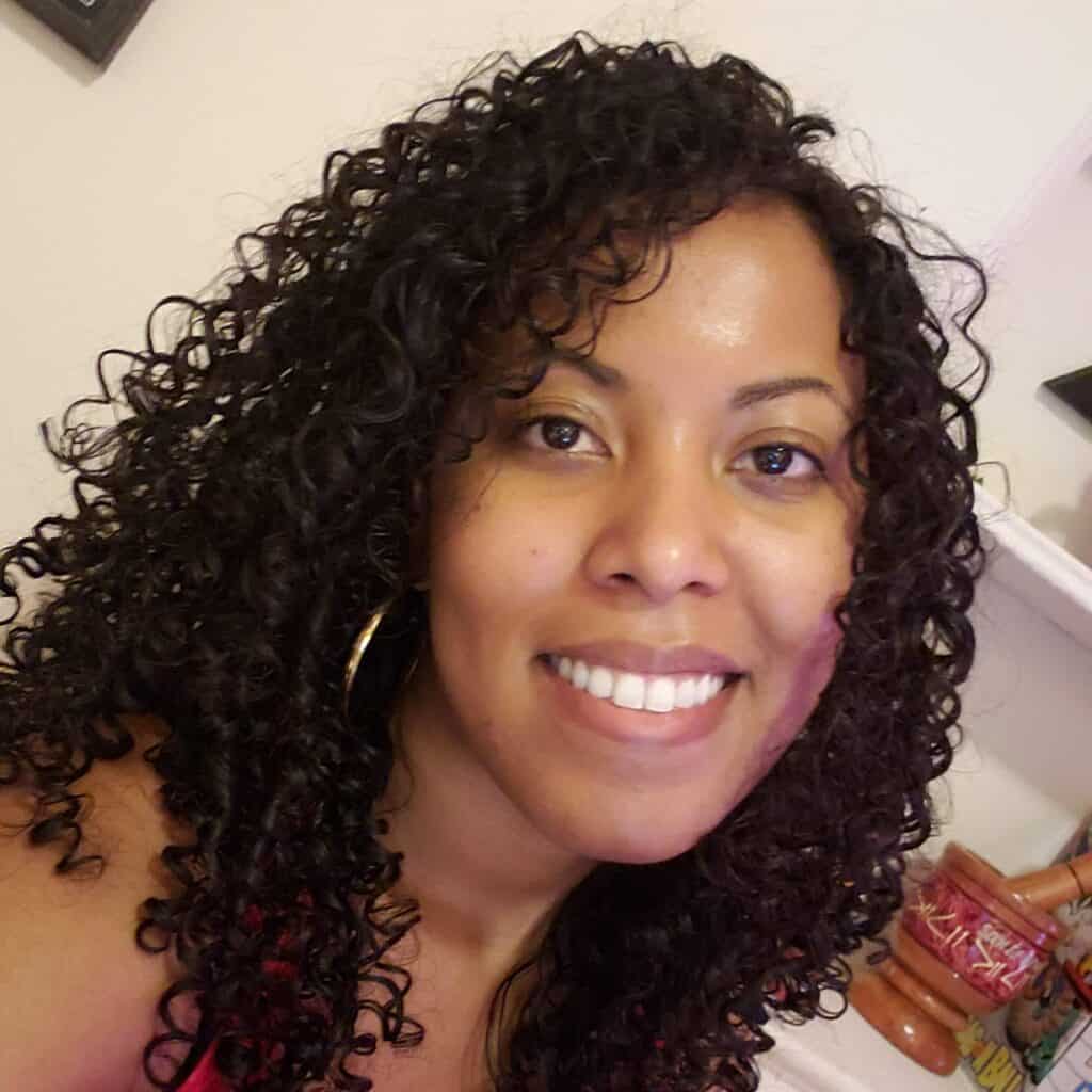 Smiling woman with long, defined black curls standing in a sunlit room, showcasing healthy, voluminous hair.