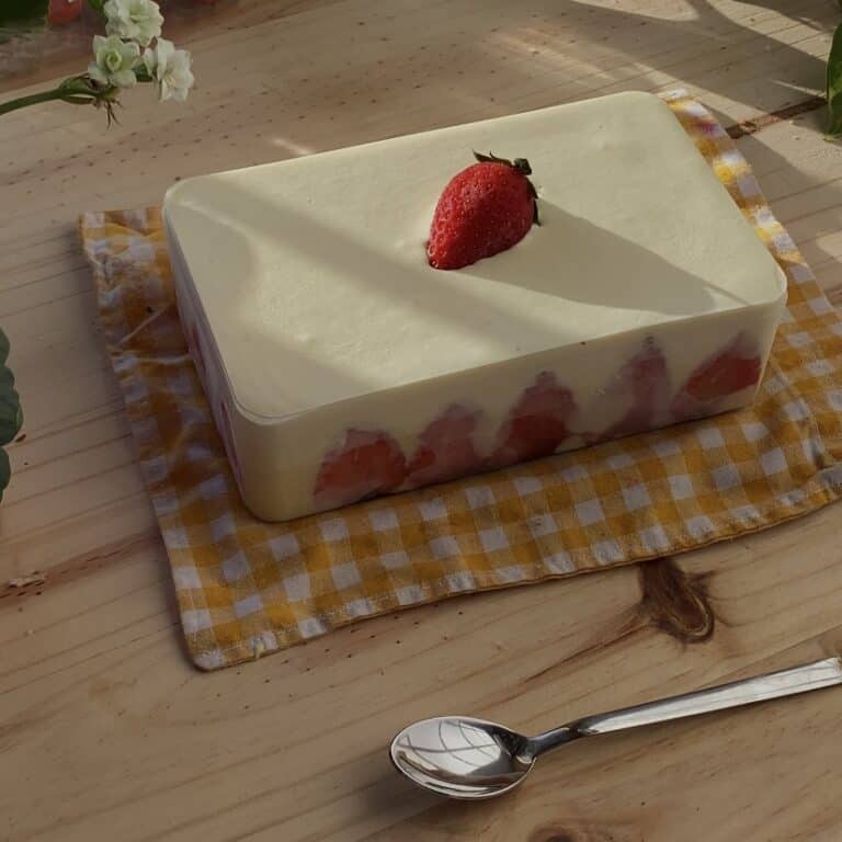 A close-up of a strawberry tiramisu served in a glass dish, showcasing its smooth exterior. The dessert is topped with a fresh strawberry. The soft, creamy texture of the tiramisu is visible along the edges, with a delicate contrast between the pale beige dessert and the bright red strawberry. The overall presentation is elegant and inviting.