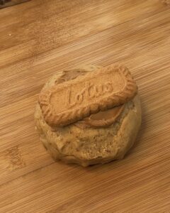 Image of a single-serve Biscoff stuffed cookie shaped into a round ball on a wooden board before baking and freezing, topped with a dollop of Biscoff spread and a Biscoff cookie for extra flavor.