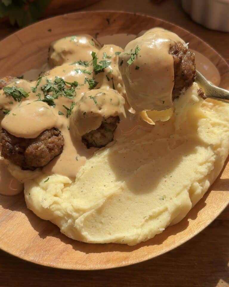 A wooden plate featuring juicy homemade meatballs covered in creamy gravy, served alongside buttery mashed potatoes, garnished with freshly chopped parsley for a flavorful and visually appealing dish.