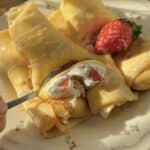 A fork cutting into a strawberries and cream crepe roll, revealing the creamy whipped filling and fresh strawberry slices inside.