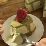 Slice of no bake pistachio cheesecake on a plate with visible crust, creamy filling, and pistachio topping, being scooped with a spoon to highlight texture