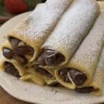 Stack of Nutella crepe rolls on a plate with Nutella oozing out and powdered sugar on top, ready to serve