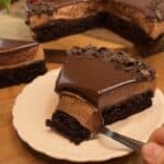 A spoon scooping the second bite of a slice of milk chocolate mousse cake, with a chocolate cake base, milk chocolate mousse filling, ganache topping, chocolate chips, fresh raspberries, and mint leaves.