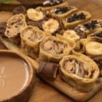 Sushi crepes on a wooden table with three varieties: Kinder Bueno spread and crushed Kinder Buenos, Nutella and banana with Ferrero Rocher, and whipped cream with chocolate and crushed Oreos, served with a bowl of Kinder Bueno spread for dipping