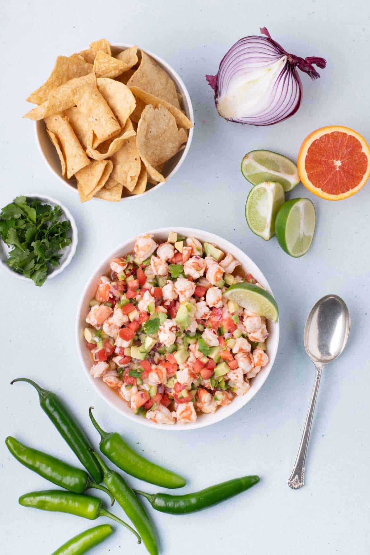 Overhead view of a bowl of shrimp ceviche surrounded by fresh ingredients like red onion, lime, orange, cilantro, green chilies, and tortilla chips, with a spoon placed beside the bowl.