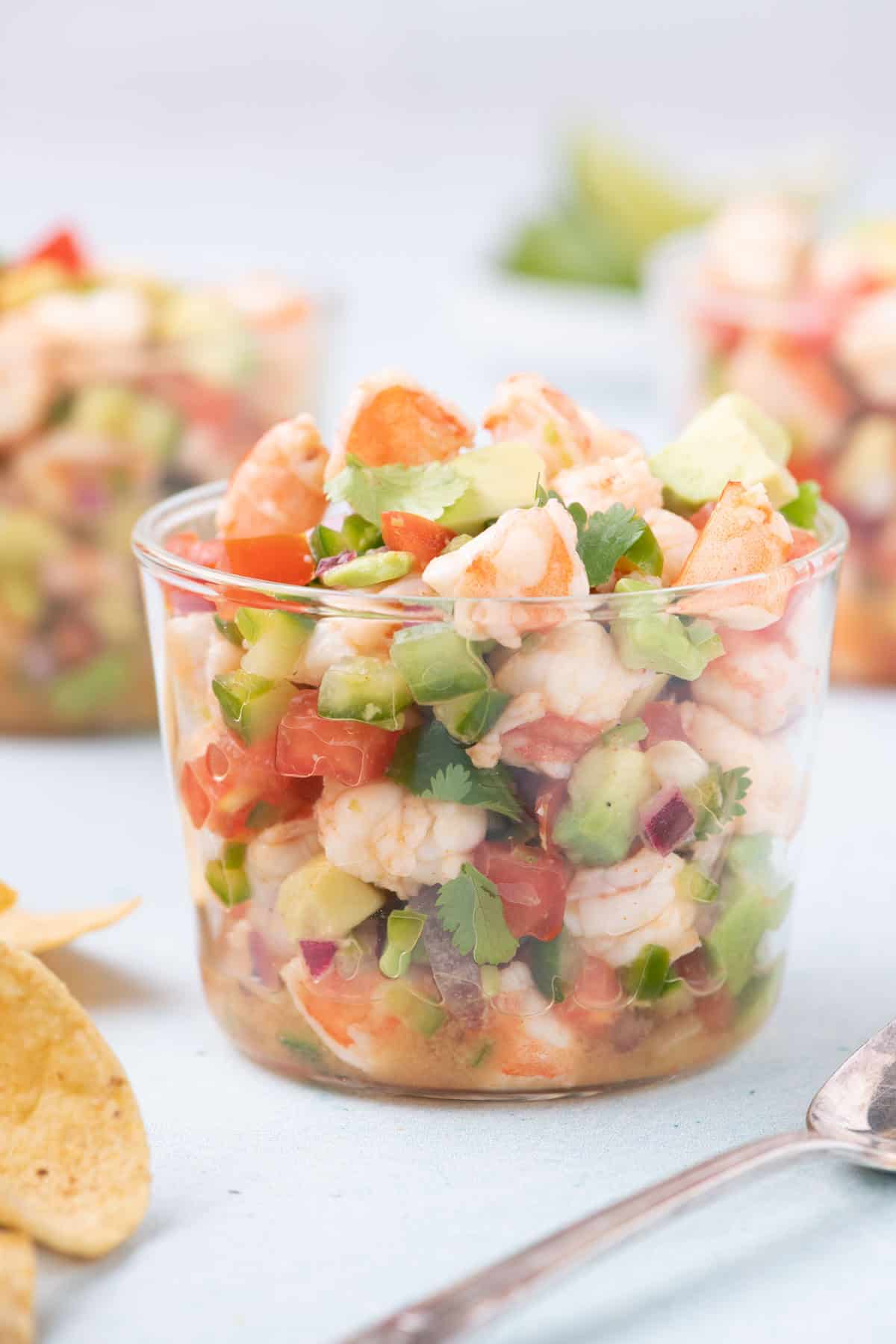 Glass filled with colorful shrimp ceviche made with avocado, tomato, jalapeño, red onion, and cilantro, with tortilla chips and a spoon nearby on a light blue surface.