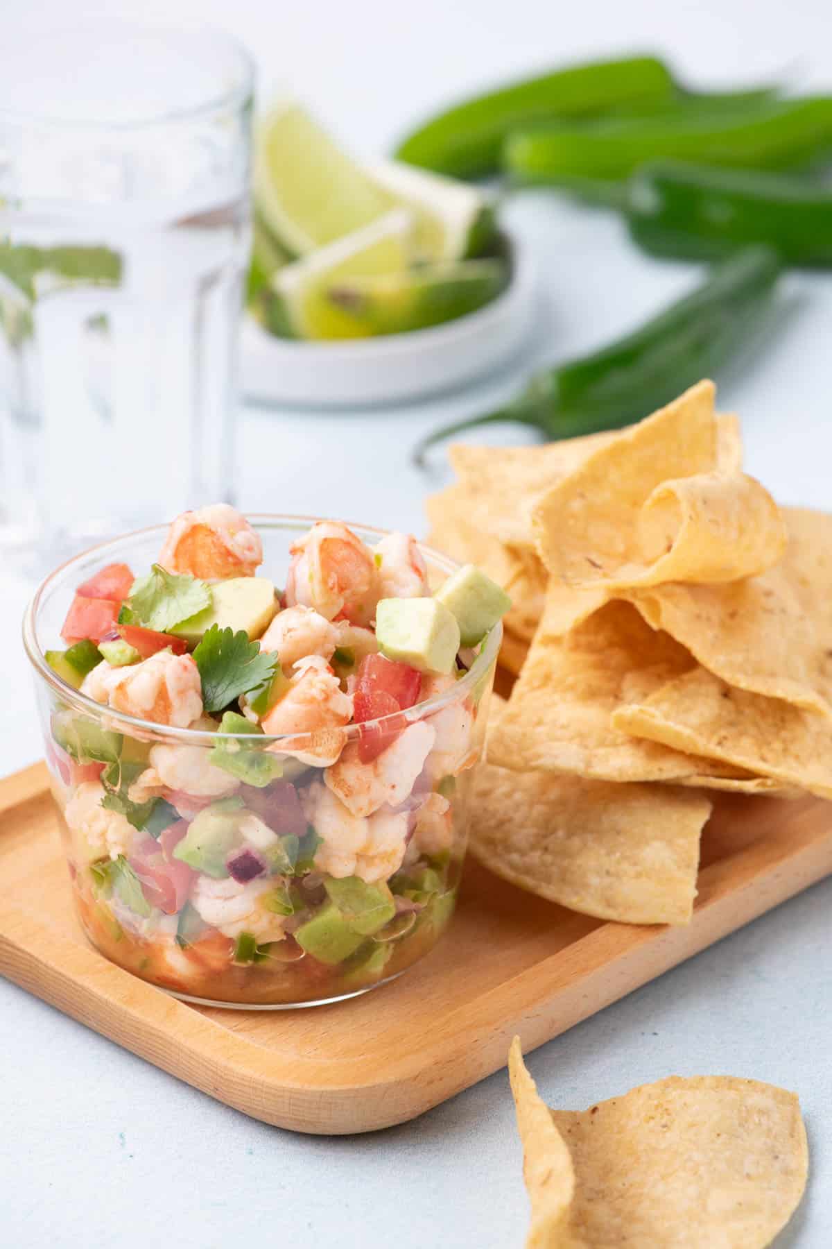 Glass cup filled with fresh shrimp ceviche made with avocado, tomato, onion, and cilantro, served on a wooden tray with crispy tortilla chips and lime wedges in the background.