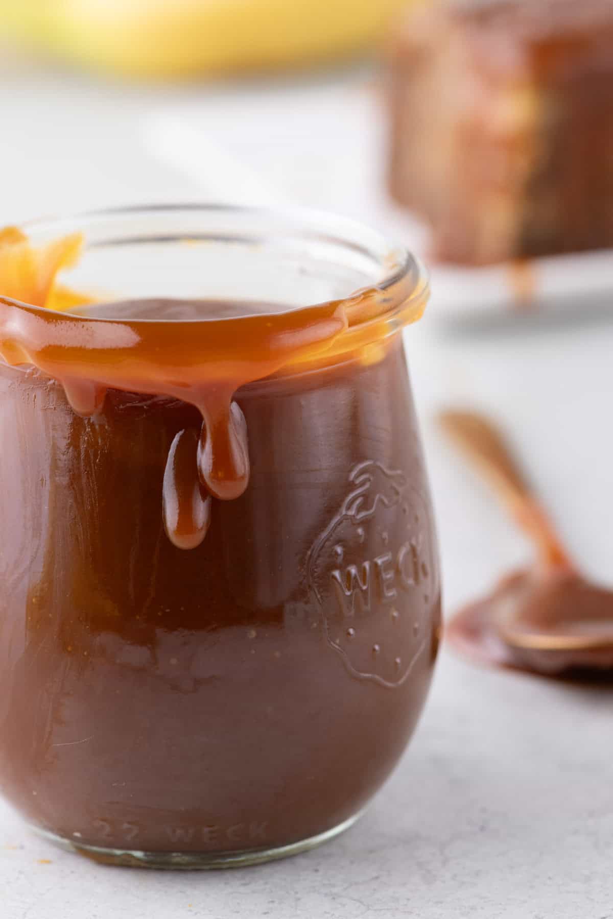 Close-up of a jar of salted caramel sauce with spoon next to a slice of banana cake drizzled with caramel