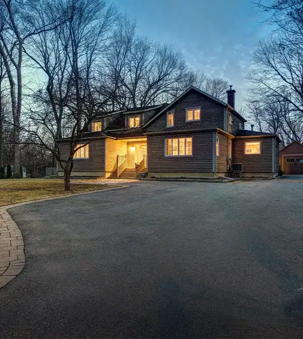 Cozy suburban house with warm interior lighting, set in a wooded area during early evening, showcasing a large driveway and classic architectural design.