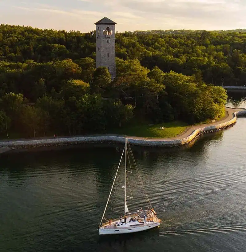 Seaside lighthouse overlooking lush green trees, sailboat on calm water near rocky shoreline, scenic coastal landscape in Atlantic destinations, perfect for boating and outdoor exploration.