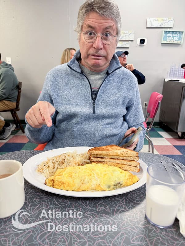 A man with gray hair and glasses looks surprised while sitting at a table with a breakfast plate including an omelet, toast, and sausage.