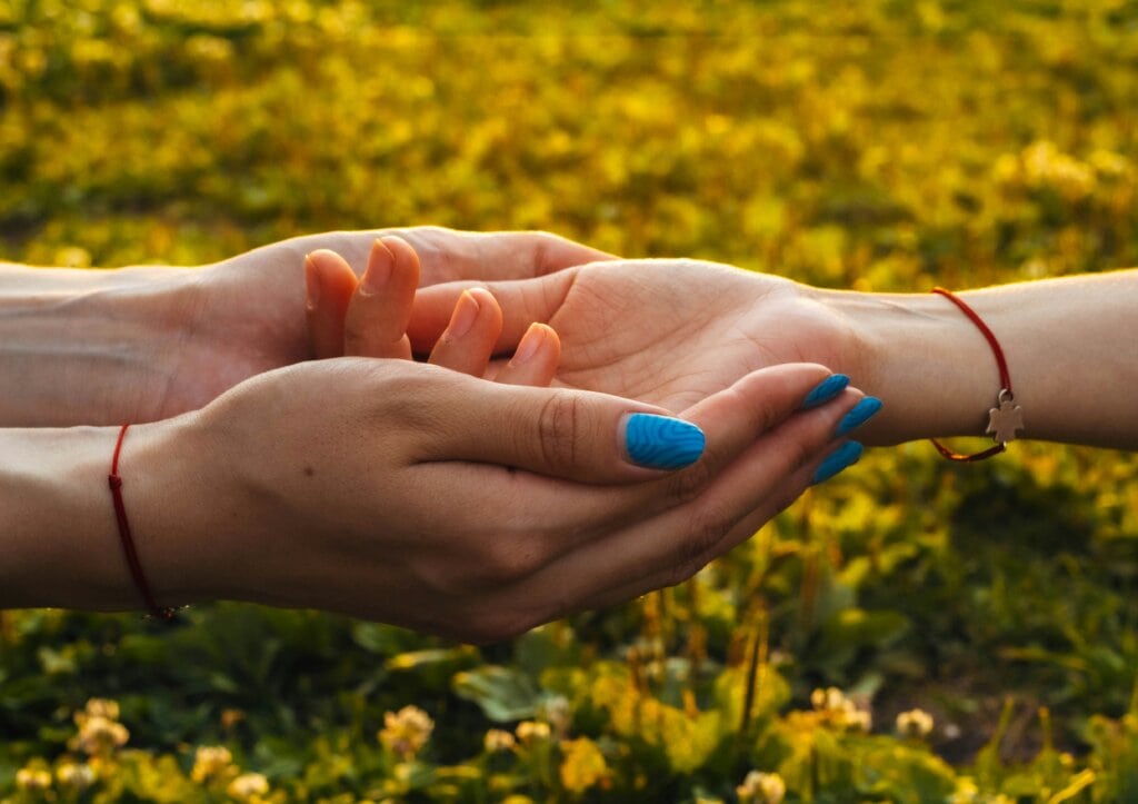 Due mani che si toccano delicatamente all'aperto con sfondo di fiori gialli. Immagine evocativa del gesto di aiuto e cura del dono, il legame invisibile ma reale che si crea tra donatore e ricevente, anche quando non si conoscono. Supporto, altruismo su un piano di parità e comunità.