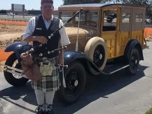 Man in Scottish attire holding a rifle next to a vintage car.