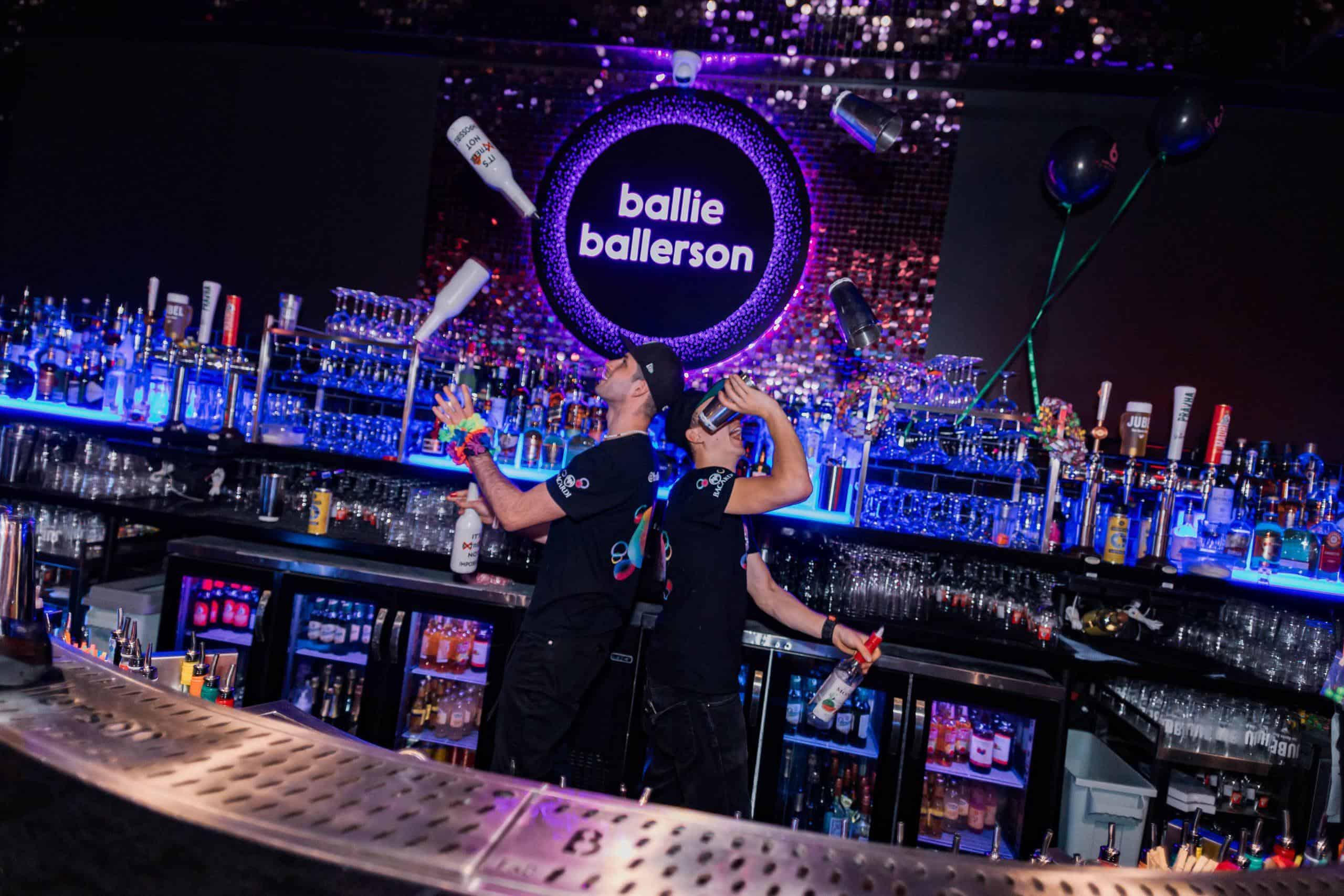 Bright, lively bar scene at Ballie Ballerson with bartenders in black shirts, colourful accessories, neon sign, and drinks.