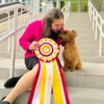 Woman with dog and large award ribbon on steps.