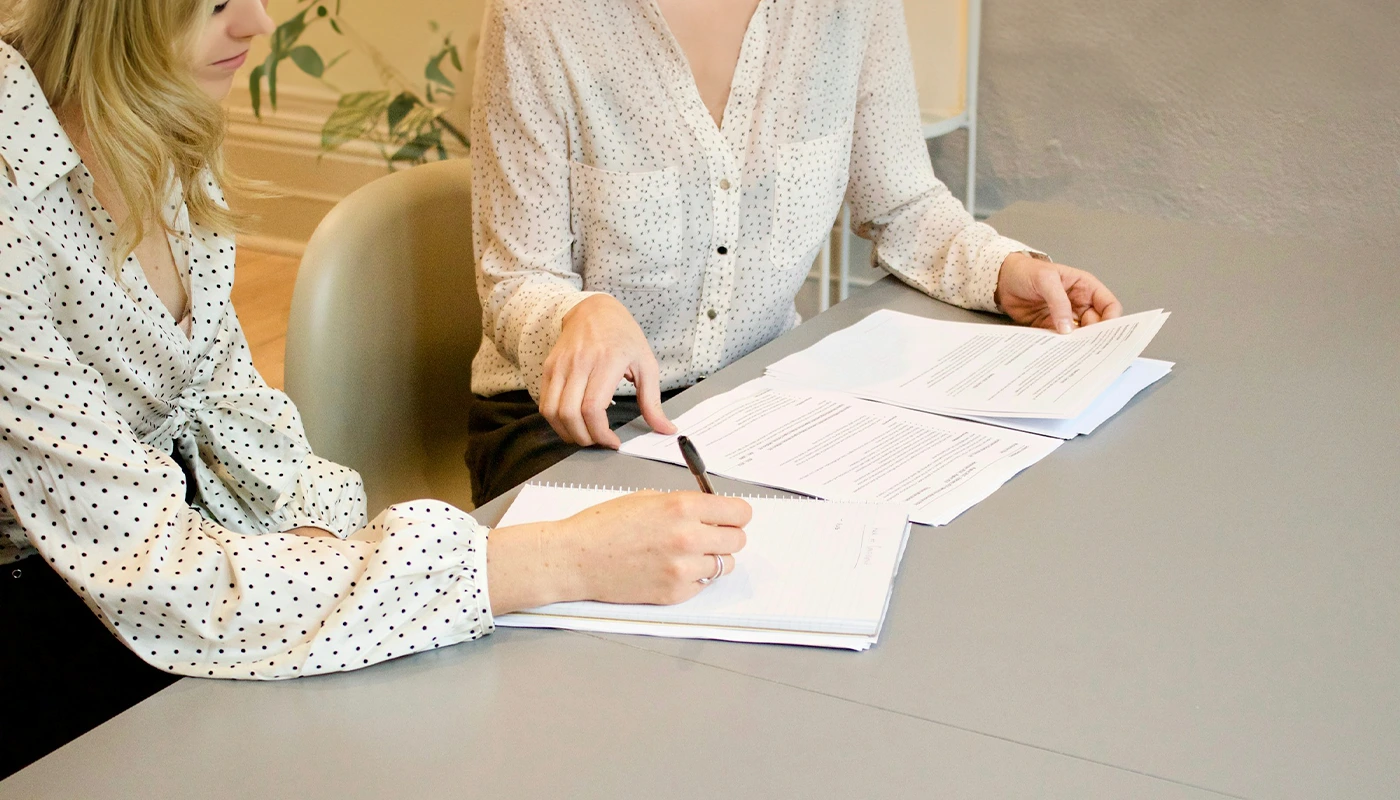 Duas mulheres analisam documentos lado a lado, com caneta e blocos de anotação sobre a mesa.