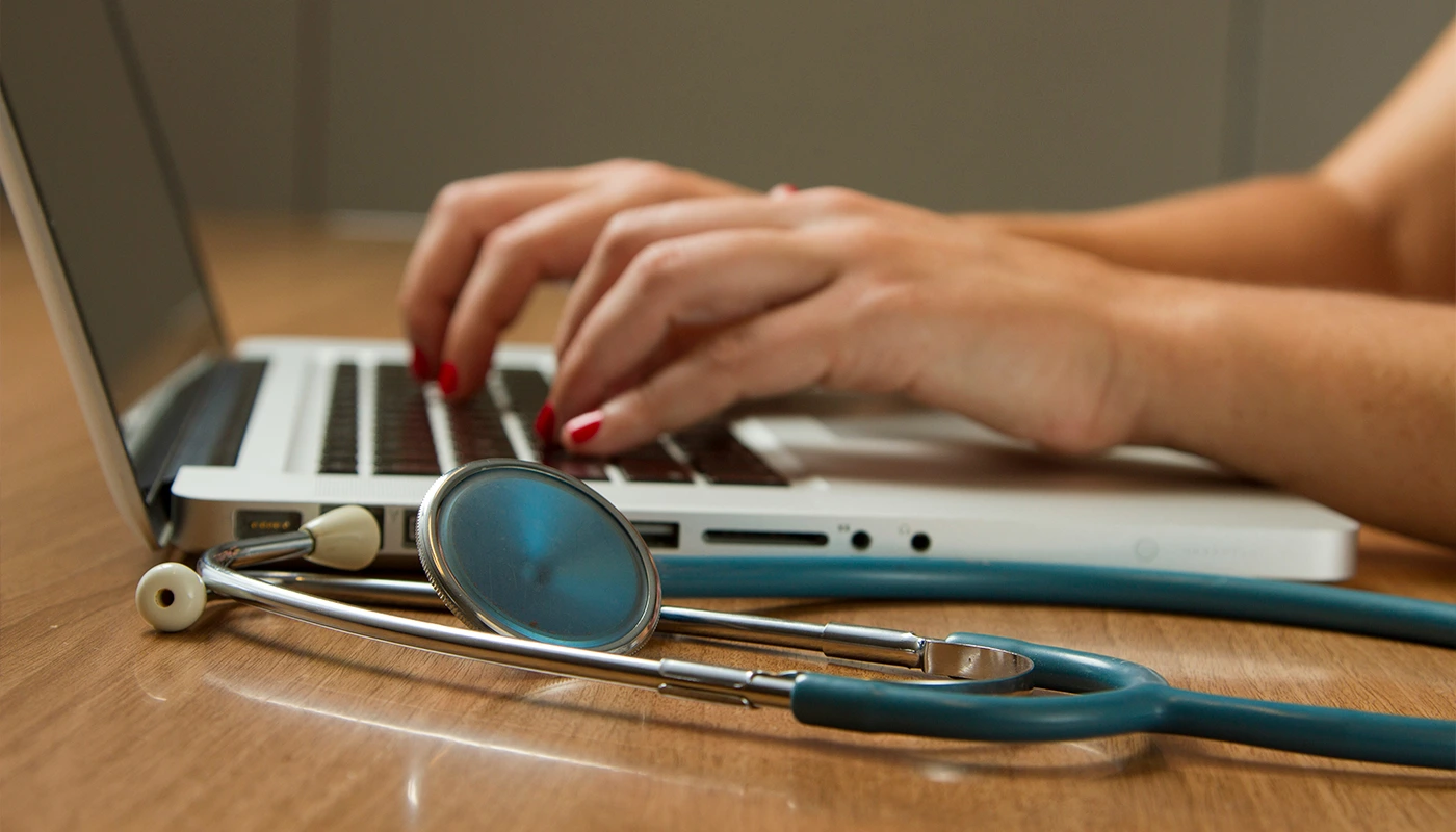 Mãos femininas com unhas vermelhas digitando em notebook com estetoscópio sobre a mesa.