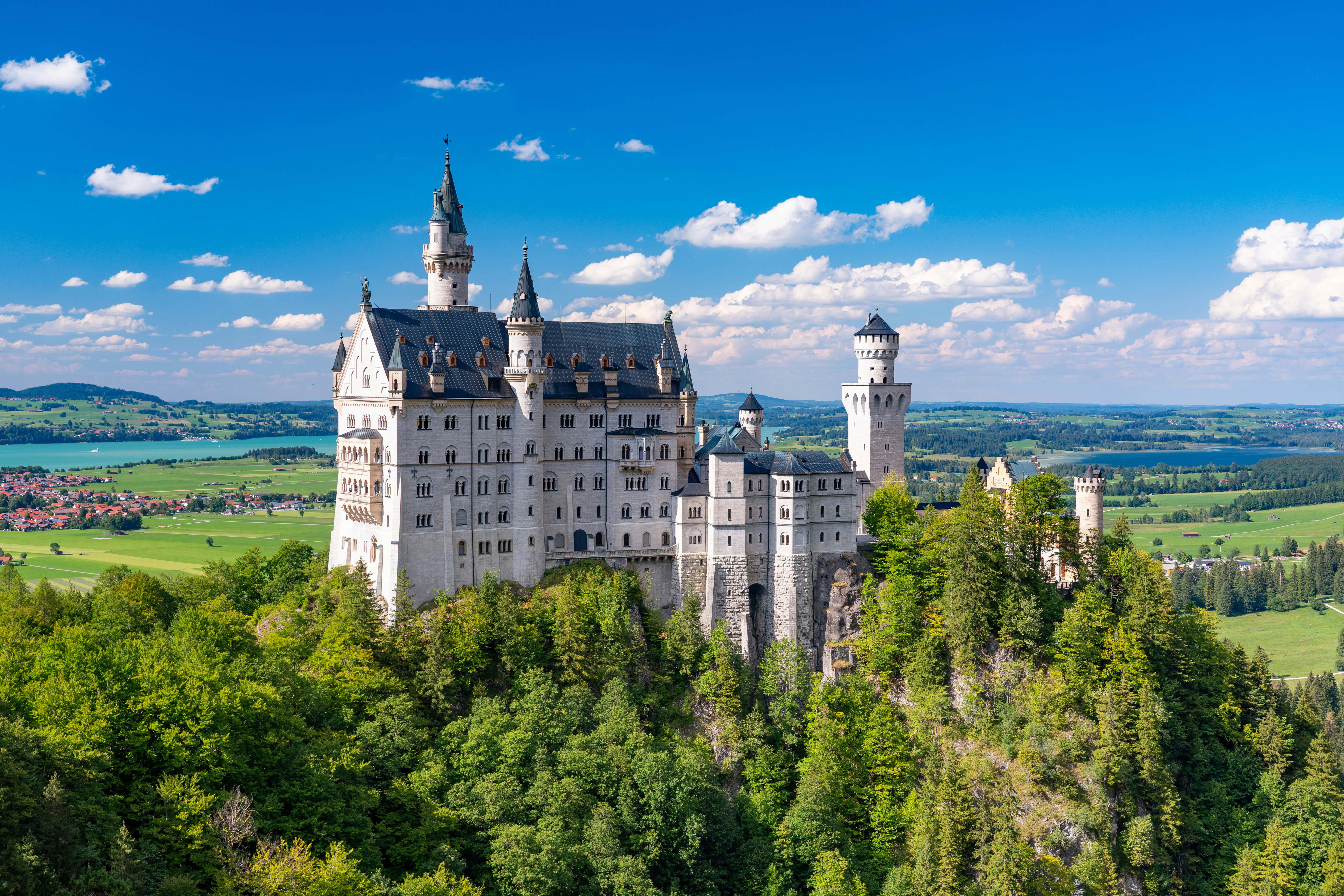 Scenic view of Neuschwanstein Castle surrounded by lush green forests and rolling hills under a bright blue sky.