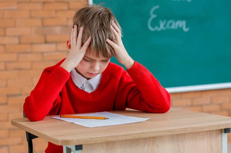 The image shows a young boy wearing a red sweater over a white collared shirt in a classroom setting. He is sitting at a desk with his hands on his head, looking down at a sheet of paper on the desk in front of him, with a pencil lying beside the paper. He appears to be struggling or overwhelmed which could represent the challenges faced by neurodiverse individuals during traditional testing environments.