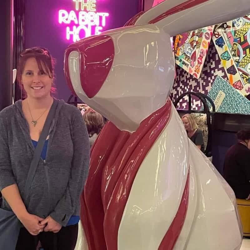 Woman in blue shirt standing next to a giant 7ft tall red and white rabbit sculpture with neon words on a wall in the back that say The Rabbit Hole.