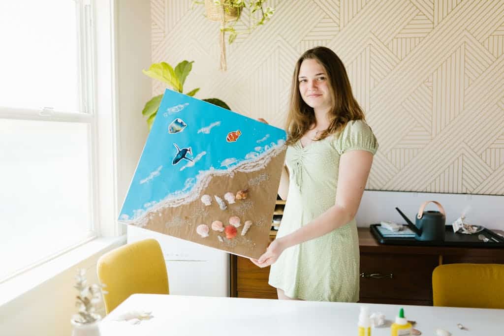Girl holding a hobby art project of the beach she created with sand, shells and sea life stickers. 