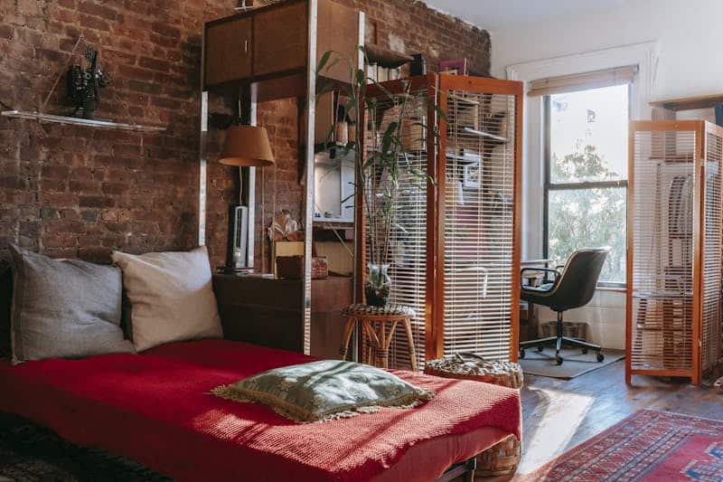 Comfortable bed with red cover and pillows placed near brick wall in cozy room with isolated workspace decorated with houseplants near window