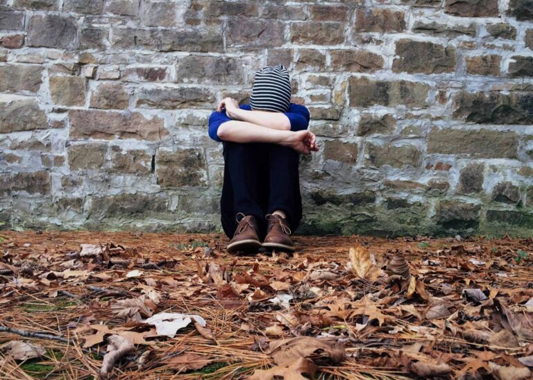 A solitary figure sits against a stone wall surrounded by fallen leaves, expressing isolation and its effects on emotional health.