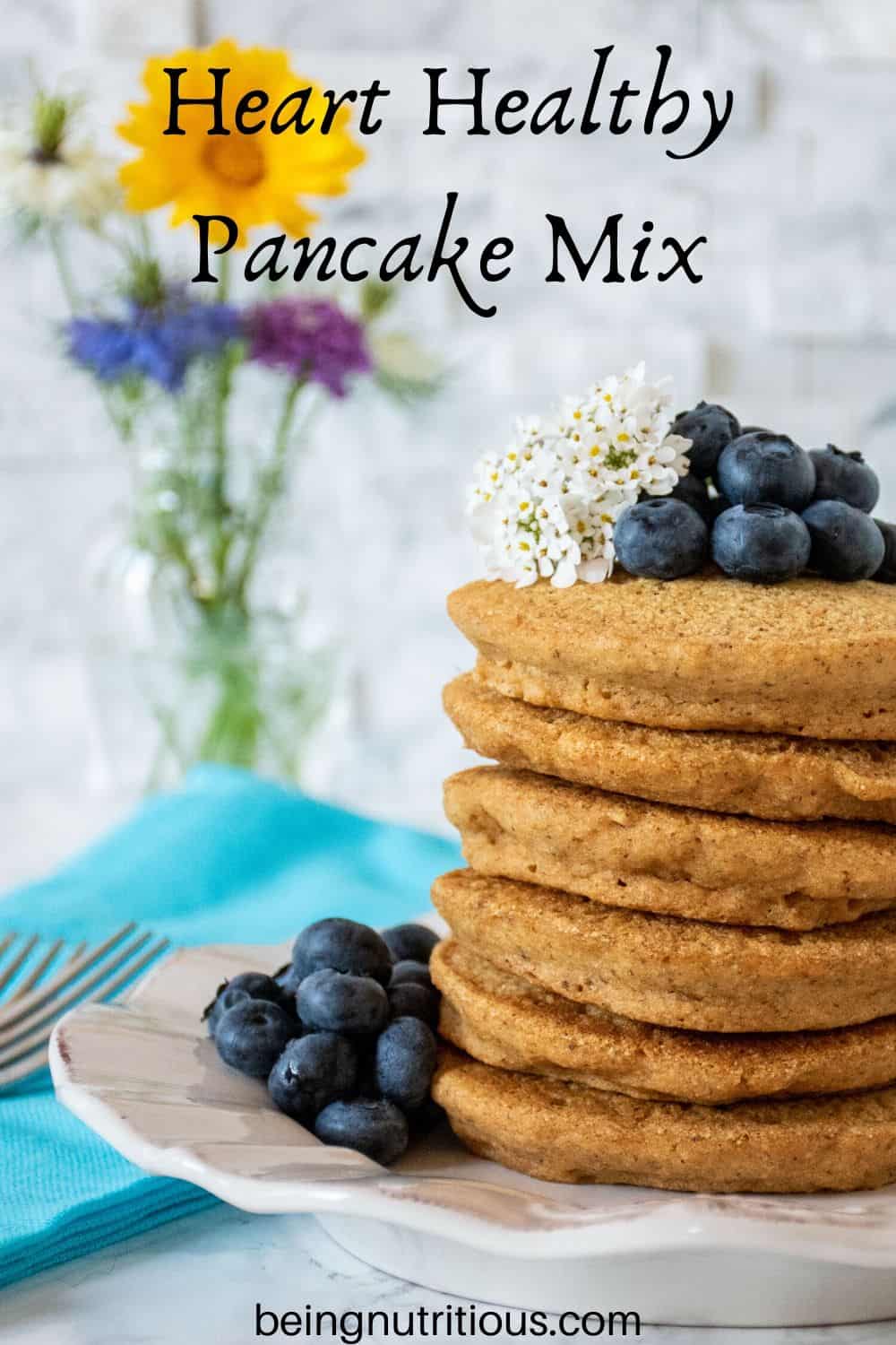 Stack of 6 whole wheat pancakes, with a pile of fresh blueberries and white flowers on top. A small bouquet of wildflowers is visible in the background.