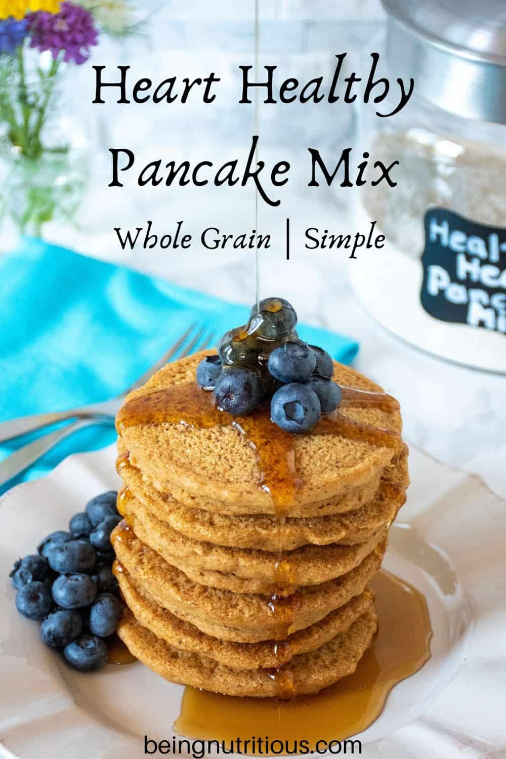 Stack of 6 whole wheat pancakes, with a pile of fresh blueberries on top, and syrup being poured over. Glass jar of heart healthy pancake mix is visible in the background.