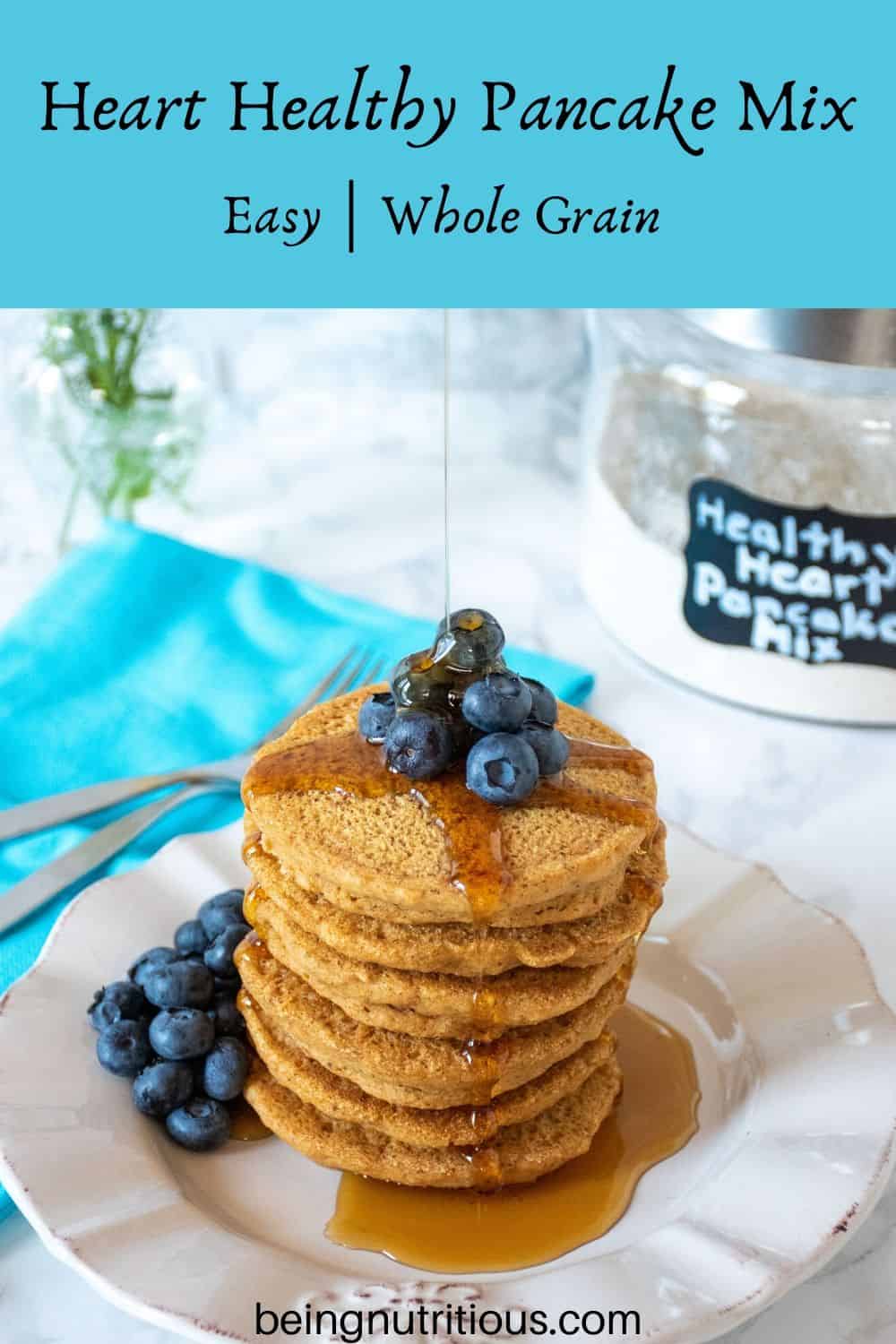 Stack of 6 whole wheat pancakes, with a pile of fresh blueberries on top, and syrup being poured over. Glass jar that reads 'healthy heart pancake mix' is visible in the background.