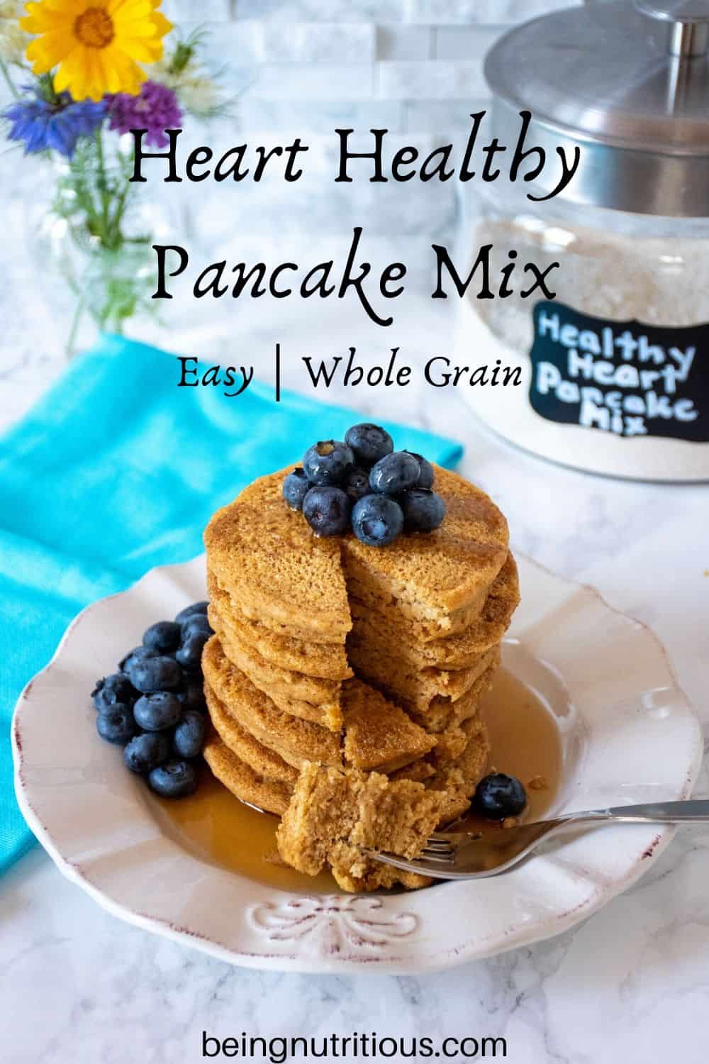 Stack of 6 whole wheat pancakes, with a pile of fresh blueberries on top, and syrup poured over. A fork with cut pieces of pancakes on it lies in front of the stack. Glass jar that reads 'healthy heart pancake mix' is visible in the background with a bouquet of flowers.