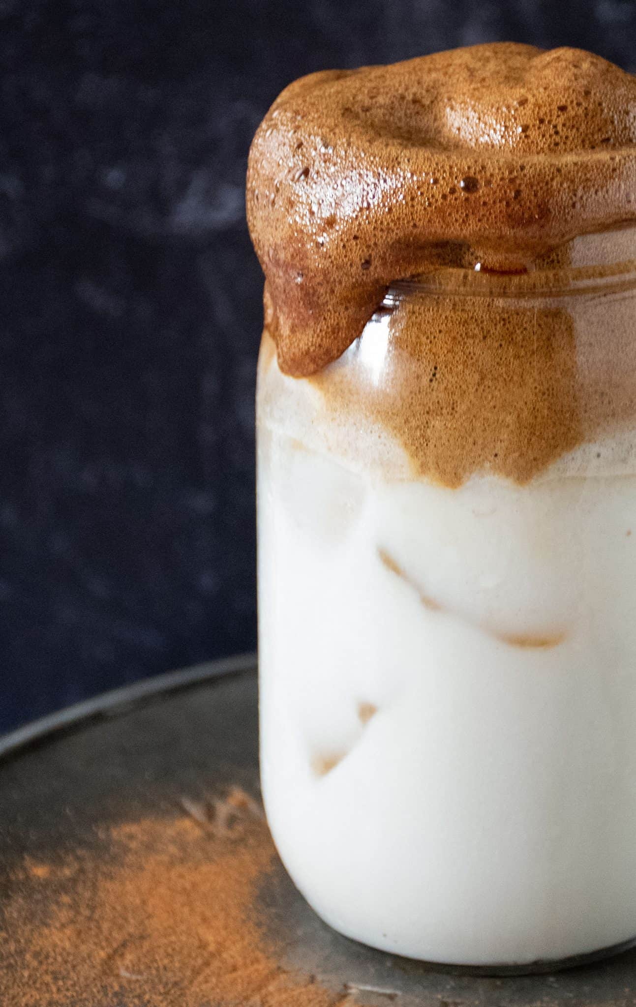 Whipped coffee, over iced almond milk, in a Mason jar in front of a dark background.