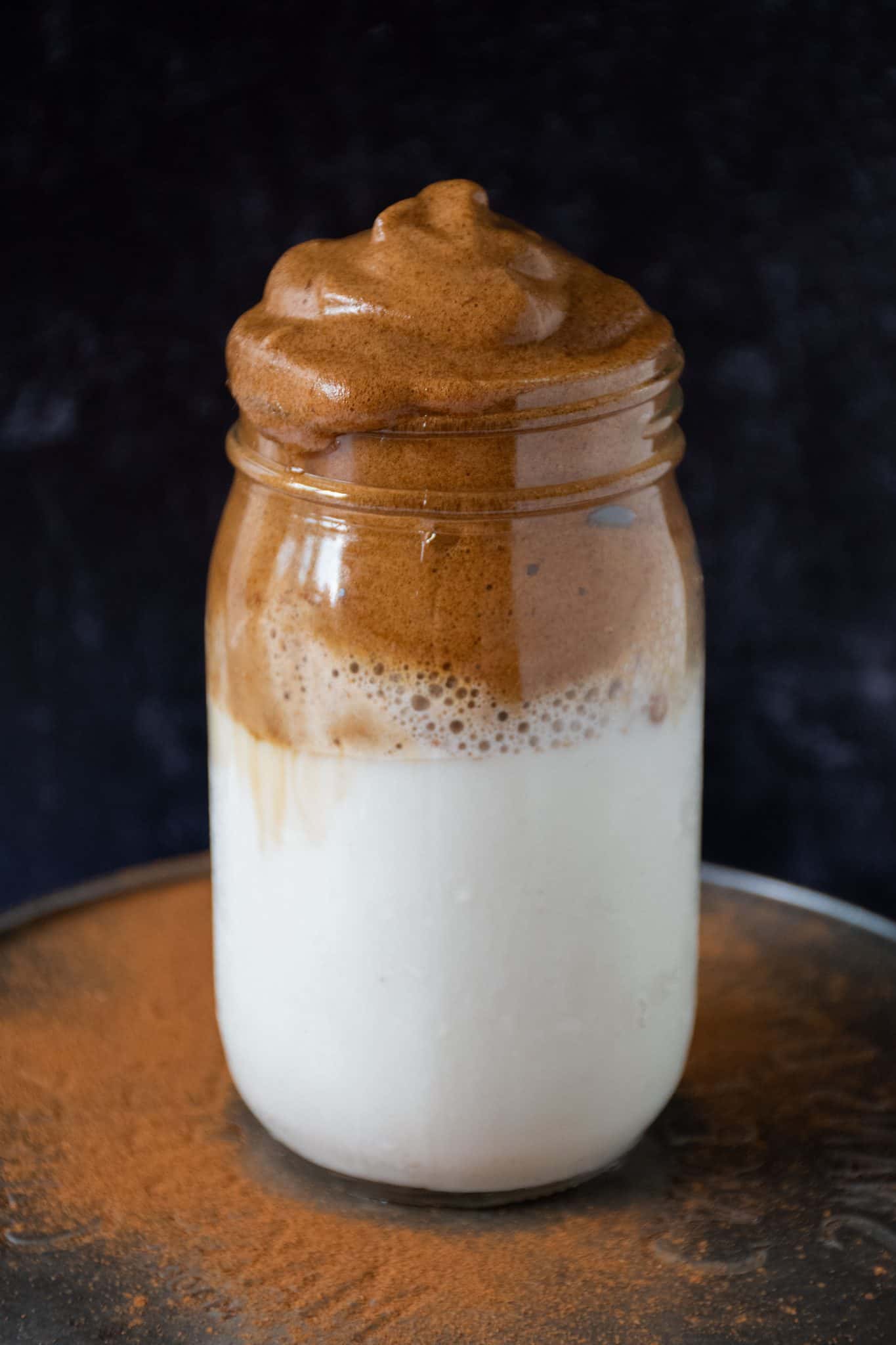 Frothy, whipped coffee, over iced almond milk, in a Mason jar in front of a dark background.