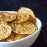 Zucchini chips in a white bowl in front of dark background.